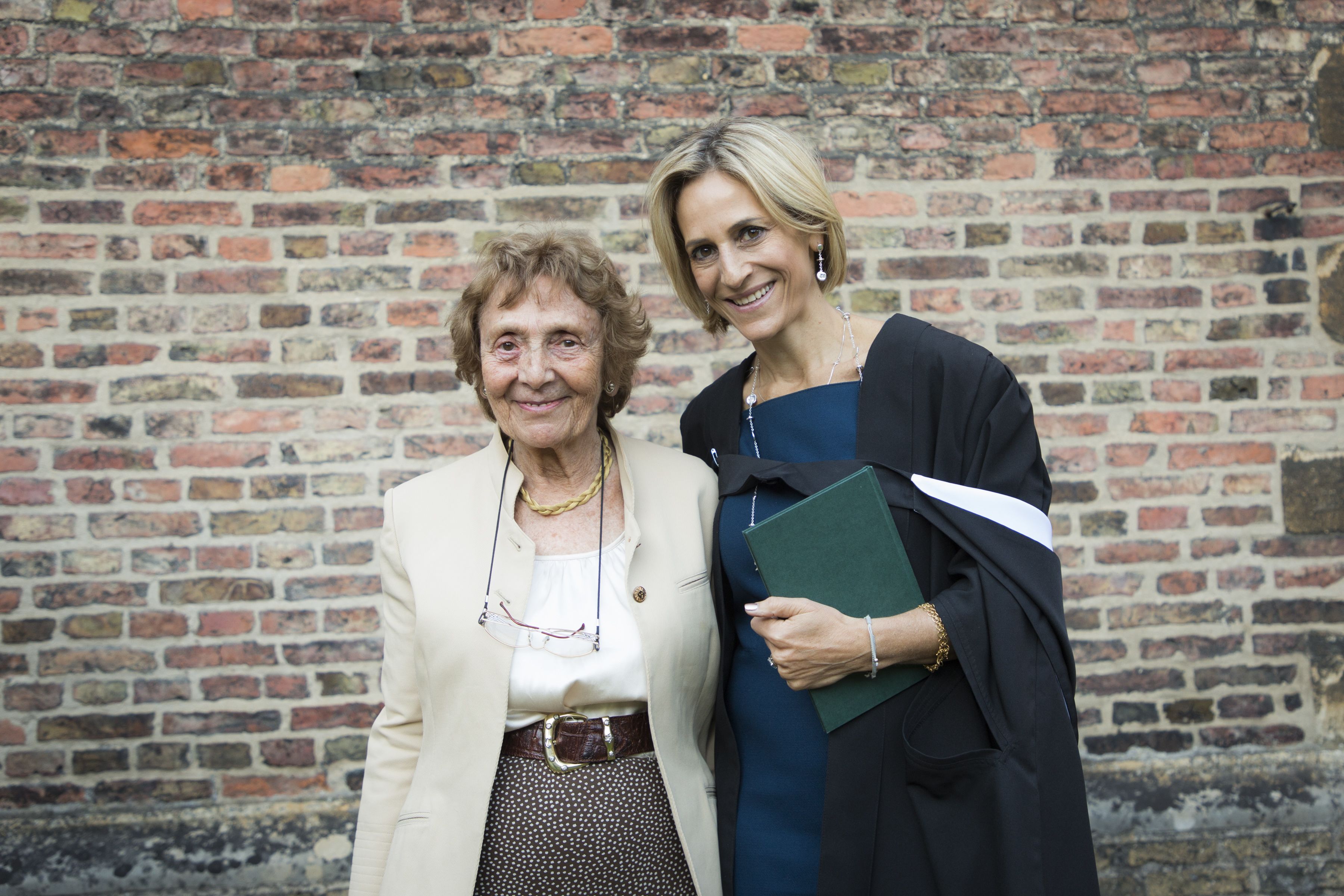 Emily Maitlis posing for a photo with her mother at the Honorary Fellows admission in 2018. Emily is holding a green book and wearing a dark blue dress with a white trimmed gown over the top. Her arm is around her mother, who is wearing a beige jacket and patterned trousers.
