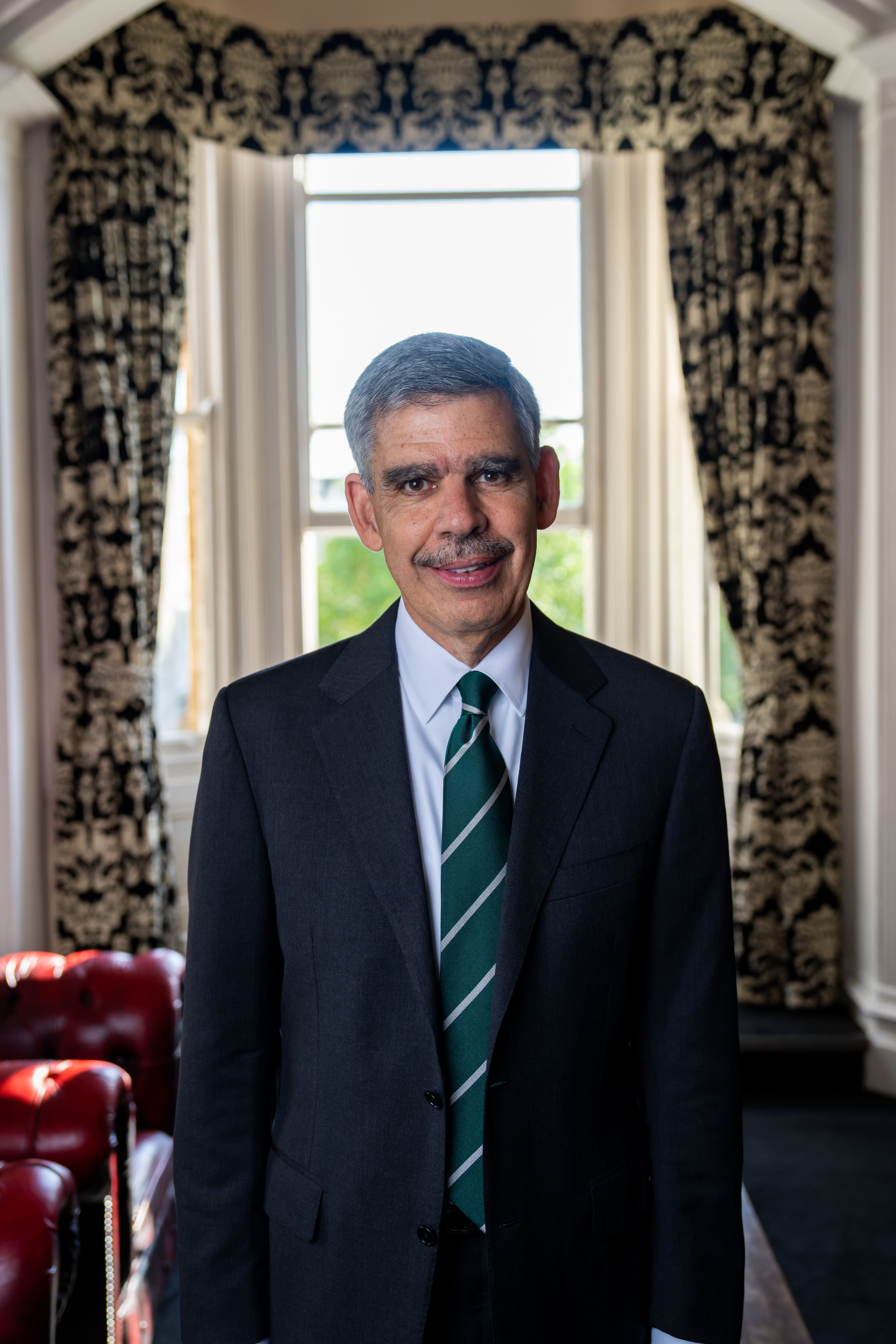 The President, Dr Mohamed El-Erian, looking at the camera in a room at the Cambridge Union with ornately patterned curtains. The President wears a suit and a dark green tie.