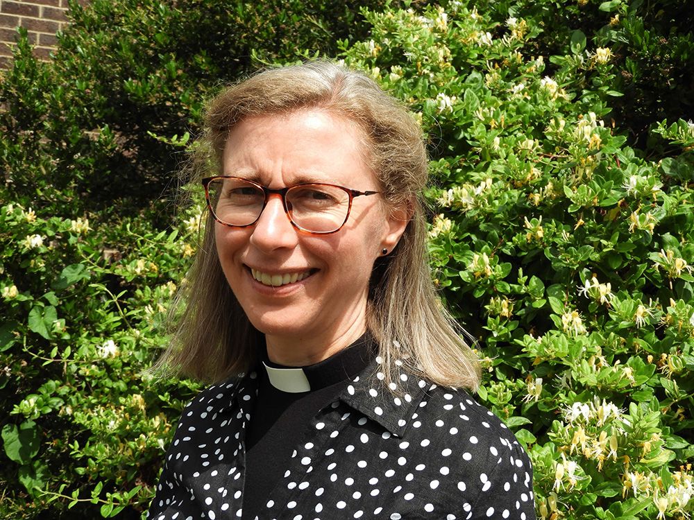 A female vicar wearing a dog collar standing in front of a hedge