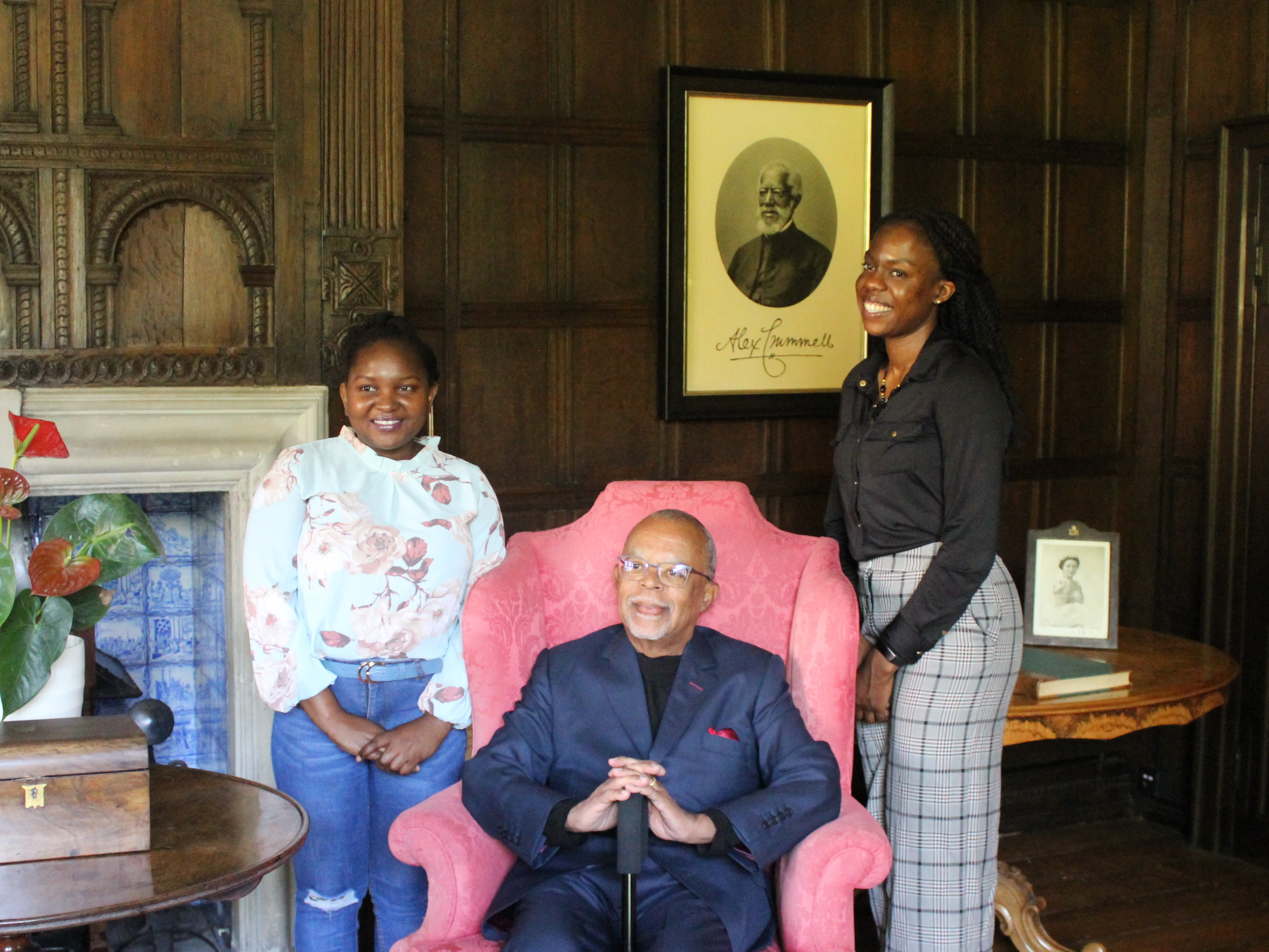 A man sat down in a pink chair with two women stood next to him