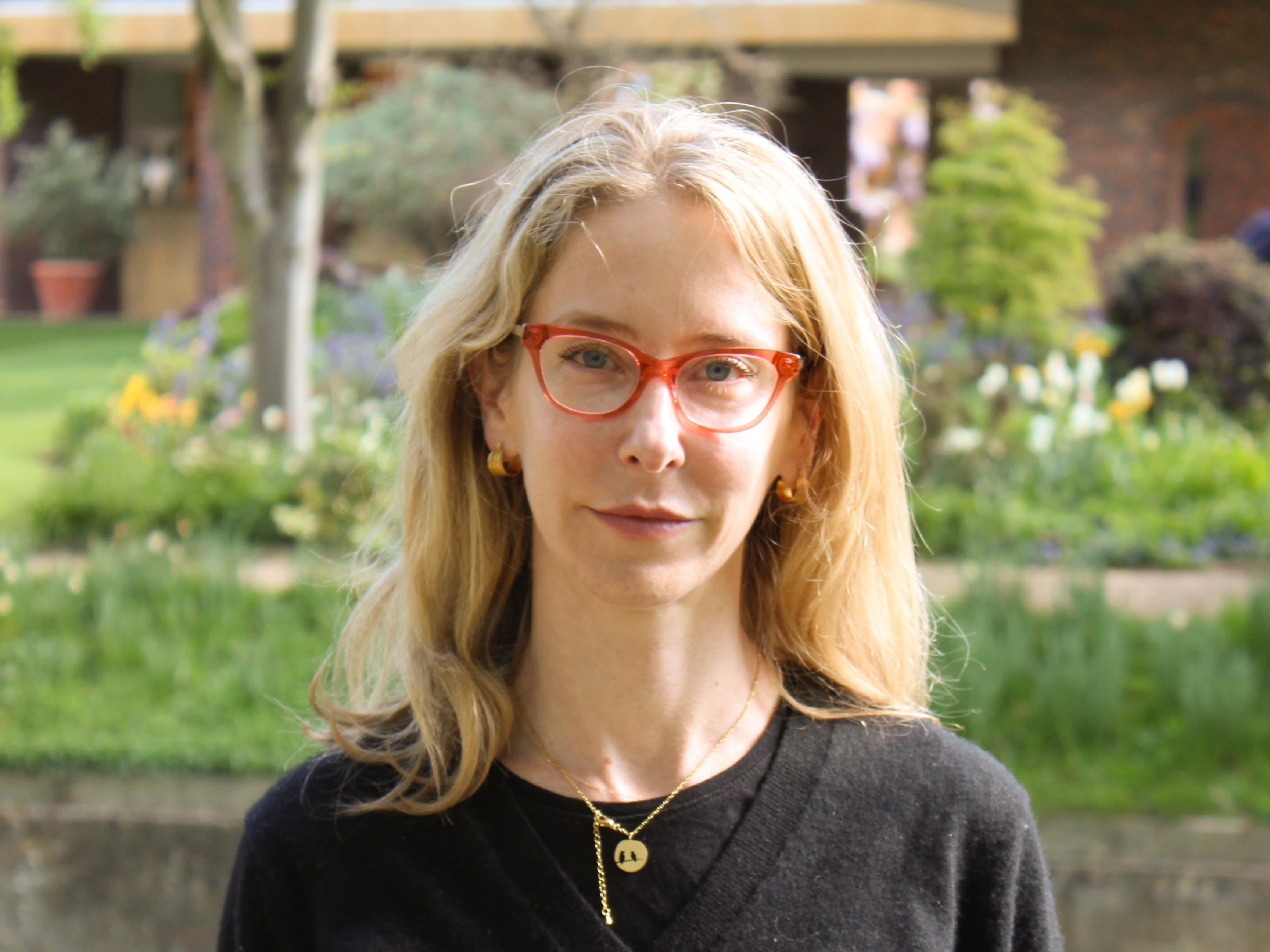 A woman with blonde hair and red glasses in front of a garden