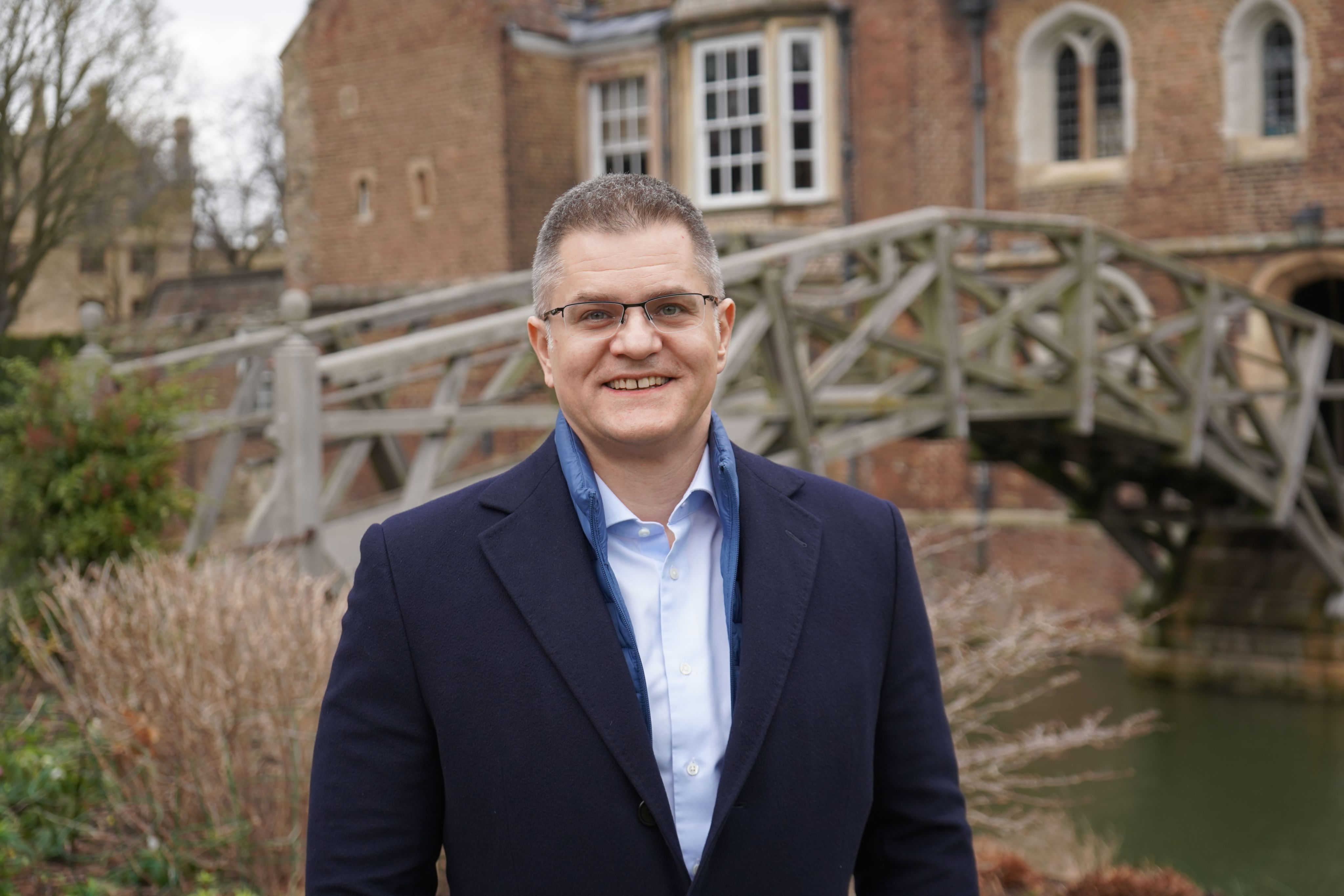 Vuk Jeremić, a man wearing a black coat over a blue jacket and a white shirt, stands in front of the Mathematical Bridge and smiles. He is wearing glasses and has cropped grey hair.