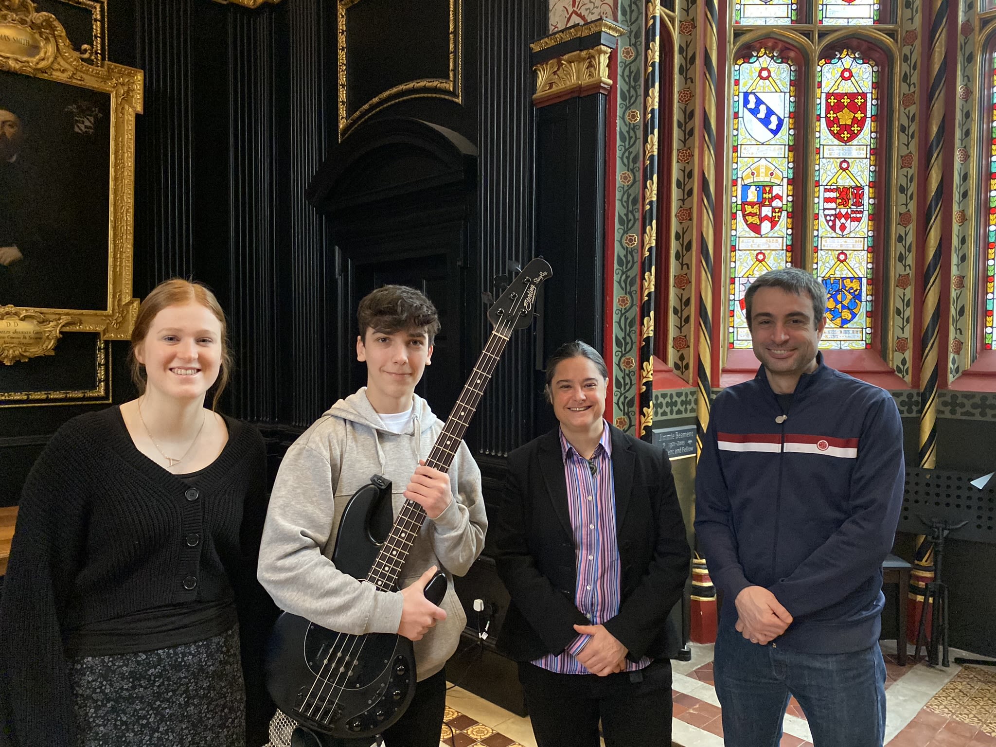 Four people lined up in Old Hall for a photo, one holding an electric bass guitar